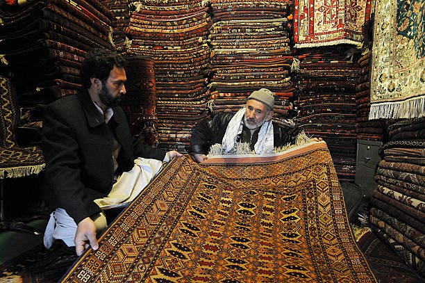 An Afghan shopkeeper (L) displays a carpet to a customer in his shop on Chicken Street in Kabul on February 26, 2008. Afghan carpets, made mostly in the north of the country, are one of the war-ravaged country's few exports and can fetch thousands of dollars each on the international market. AFP PHOTO/SHAH Marai (Photo credit should read SHAH MARAI/AFP via Getty Images)